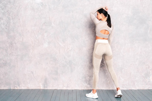 Back view of fitness confident woman in sports clothing Sexy young beautiful model with perfect body Female posing near grey wall in studio Stretching out before training