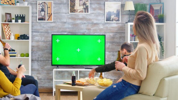 Вид сзади на друзей, пьющих пиво и смотрящих спорт по телевизору с зеленым экраном в гостиной.