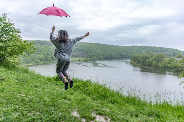 Back view of a girl under an umbrella jumping near a lake in a mountainous area in rainy weather.