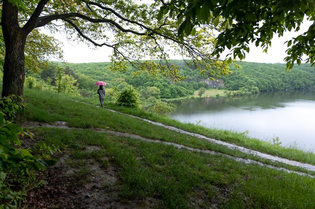 Back view of a girl under an umbrella on a walk in the forest near the lake in rainy weather.