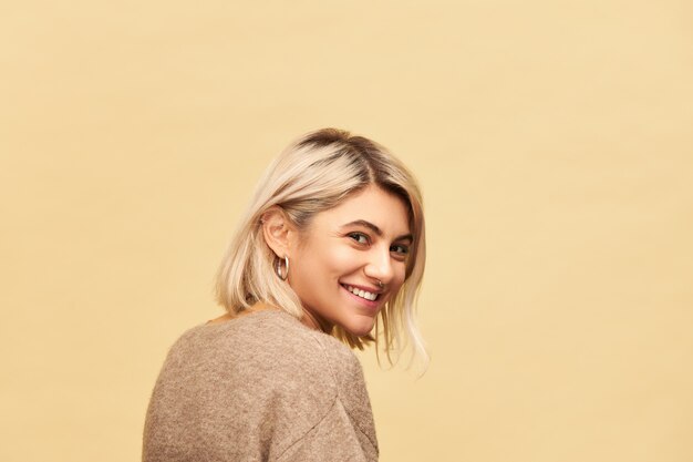 Back view of gorgeous positive young European female wearing warm cozy jumper turning head round and  with radiant happy smile, being in good mood, having playful facial expression