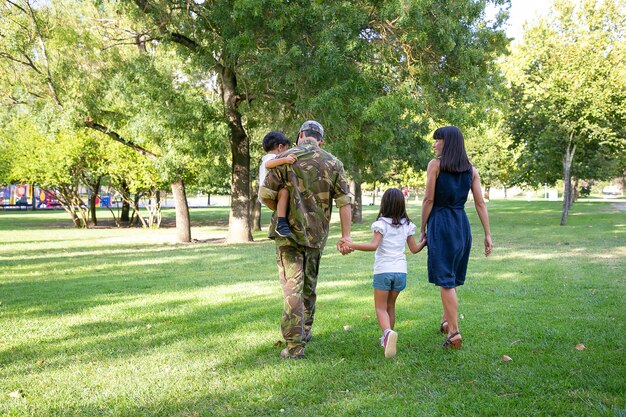 公園の牧草地で一緒に歩いている幸せな家族の背面図。迷彩服を着て息子を抱き、妻と子供たちと週末を楽しんでいる父。家族の再会と帰国のコンセプト