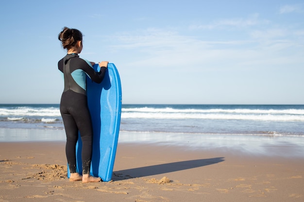 Вид сзади маленькой девочки с доской для серфинга на пляже. Темноволосый ребенок в форме для серфинга в солнечный день смотрит на воду. Спорт, отдых, концепция активного образа жизни