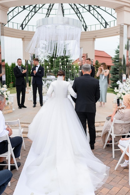 Back view of loving father with bride daughter in long puffy white dress go to the goom on wedding ceremony outdoors Touching moment for guests and married couple Stylish wedding altar