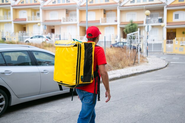 Back view of man in red cap carrying yellow thermal bag. Deliveryman working at post and delivering order on foot.