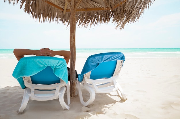 Uomo posteriore di vista che si rilassa sotto l'ombrello della palma alla spiaggia del ricorso