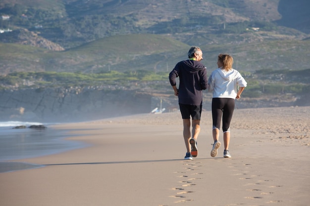 Back view of mature couple jogging on sea beach. Caucasian man and woman wearing sportswear training strengthening health and enjoying time together. Health, active lifestyle of aged people concept