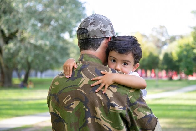 Back view of middle-aged father holding and embracing his son. Lovely little boy hugging dad in army uniform and looking away.