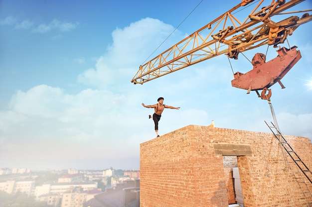 Back view of muscular and athletic man doing exercise on high brick wall. Un finishing building on high. Big iron crane and cityscape on background.