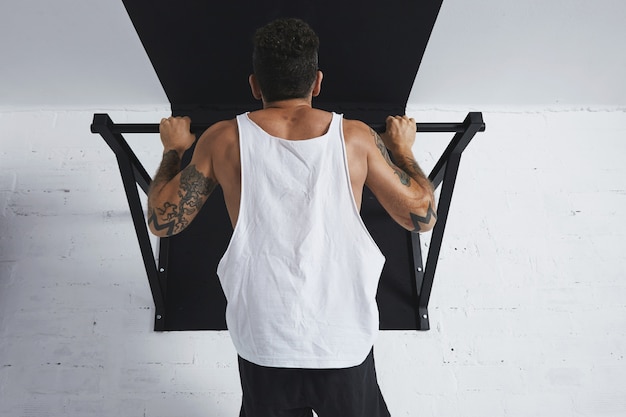 Back view on muscular male athlete in white blank tank t-shirt showing calisthenic moves Pull up on pullbar, holding on top