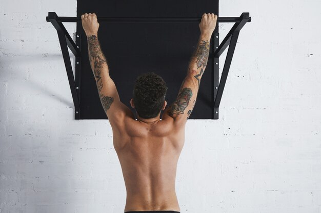 Back view on muscular topless male athlete showing calisthenic moves Hanging on pullbar