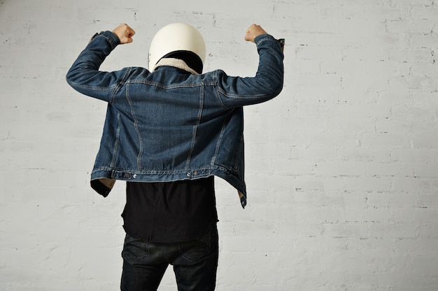 무료 사진 younf 오토바이 운전자의 몸에 맞는 뒷모습은 헬멧, 검은 색 긴 소매 헨리 셔츠 및 클럽 데님 재킷을 손으로 착용합니다.