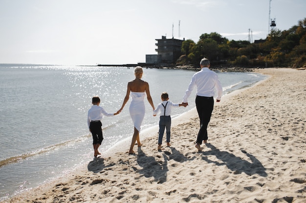 부모와 자녀의 후면보기 함께 손을 잡고 화창한 여름 날에 해변에서 걷고, 흰색 세련된 옷을 입고
