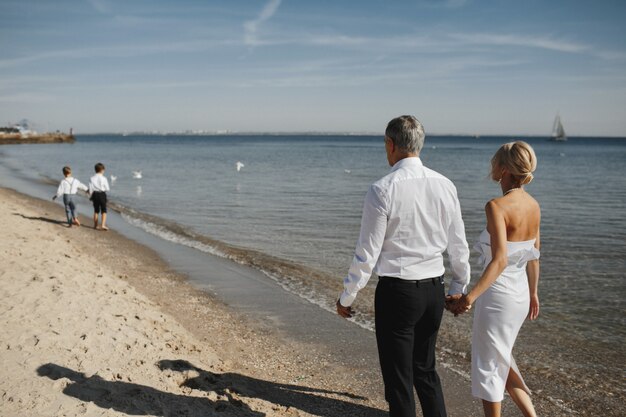 Back view of parents, who are holding hands together and two young sons in front of them on the coastline