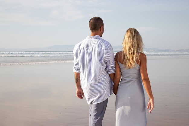 해변을 걷고 있는 행복한 커플의 뒷모습. 머리를 면도한 백인 남자와 캐주얼 드레스를 입은 여자가 손을 잡고 있습니다. 사랑, 휴가, 애정 개념