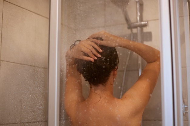 Back view portrait of slime woman with naked shoulders taking shower, posing backwards in bathroom, personal hygiene, morning procedures, hair care and refreshment.