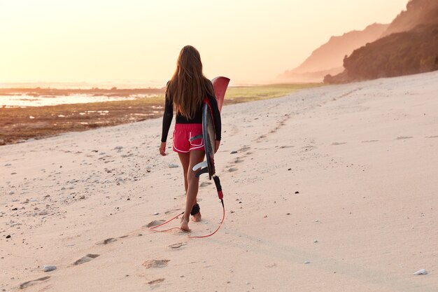 Back view of professional female surfer in diving suit walks across coastline near ocean