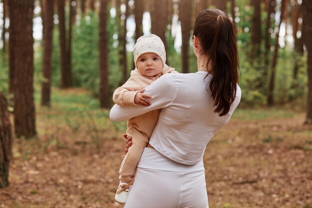 Back view of slim woman standing in forest among trees and holding infant baby in hands, kid looking at front