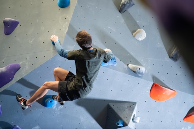 Back view of strong athlete training climbing artificial wall. Caucasian guy doing bouldering in gym holding wall with hands climbing up and balancing body. Active life and sport activity concept