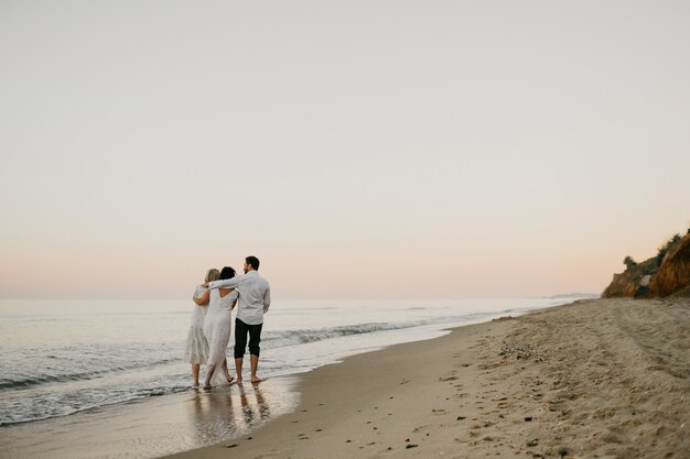 Back view of three adult humans hugging walking on the beach together