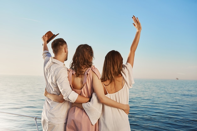 Back view of three best friends travelling by boat hugging and waving while looking at sea. People who are on luxury vacation say hi to ship crue that passes by yacht.