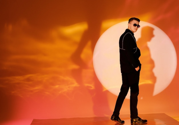 Free photo back view of trendy male in classic black suit and dark sunglasses keeping hands in trousers looking away while standing on centre of illuminated spotlight against orange background