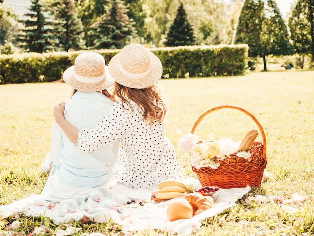 Back view of two young beautiful smiling hipster girls in trendy summer sundress and hats.Women making picnic outside.