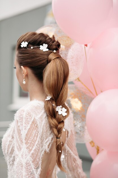 Back view of unrecognizable gorgeous brunette with beautiful hairdo and flowers in it posing in luxurious white dress with bunch on pink air balloons