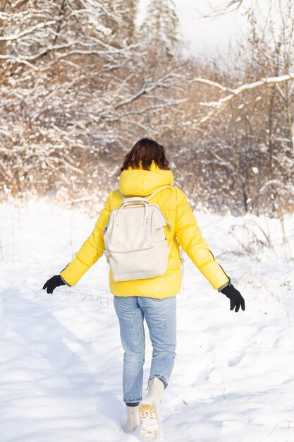 雪の風景の森の中でバックパックと明るい黄色のジャケットとジーンズの女性の背面図は雪の吹きだまりを歩きます