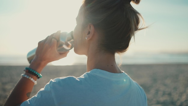 Free photo back view of woman drinking water after yoga practice on the beach sporty girl resting by the sea water balance