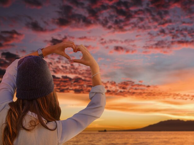 Back view woman making heart-shaped hands on a sunset