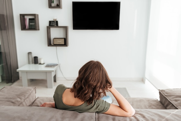 Free photo back view of woman sitting on sofa and watching tv in home