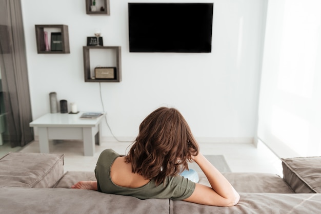 Punto di vista posteriore della donna che si siede sul sofà e che guarda tv nella casa