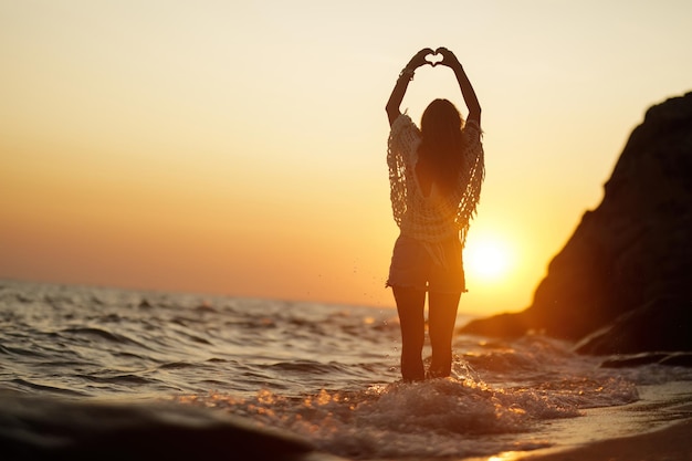 Back view of woman standing in the water and making heart shape during summer sunset Copy space