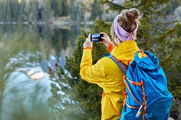 Back view of woman traveler takes photo on camera of modern cell phone, captures scenic view near mountain lake, has road trip