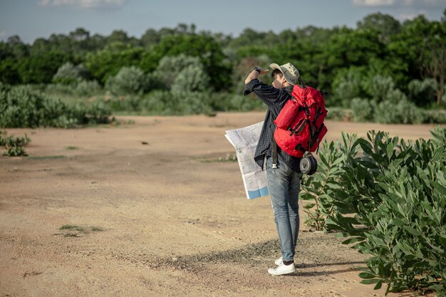 방향을 확인하기 위해 지도가 있는 모자를 쓴 젊은 백패커 남자의 뒷모습, 그는 산림 재판에서 여름 방학 동안 야외에서 휴식을 취하는 동안 큰 배낭을 메고 공간을 복사합니다.
