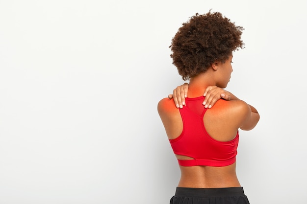 Back view of young curly woman suffers from neck pain and osteoporosis, has painful feelings in muscles, holds hands near shoulders, wears red top