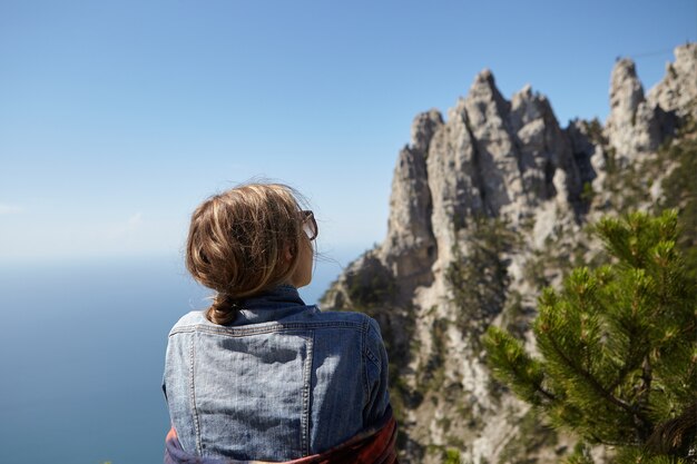 Вид сзади молодой женщины в джинсовой куртке и солнцезащитных очках, стоящей на вершине горы, любуясь великолепным морским пейзажем и панорамным видом на скалы Ай-Петри во время путешествия в одиночестве. Крымская природа.