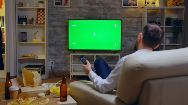 Free photo back view of young man sitting on couch in front of tv with green screen using remote control.