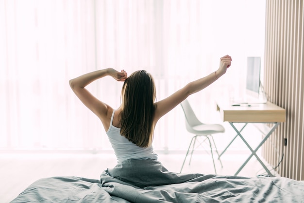 Back view of young woman stretching on unmade bed after waking up and looking at city view in the window