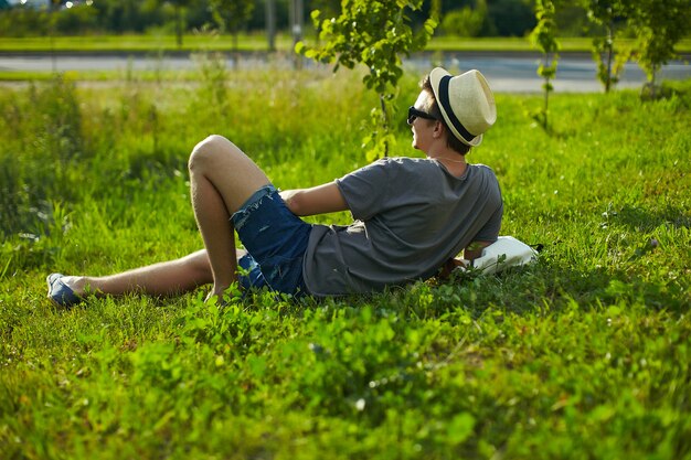 back of young attractive modern stylish man in casual cloth in hat in glasses sitting in the park in green grass
