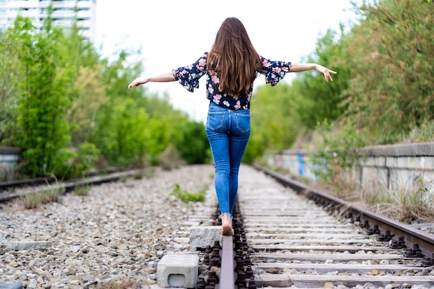 Indietro di una giovane donna che cammina attraverso i binari del treno a piedi nudi e cerca di mantenere l'equilibrio