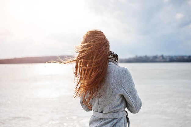 Спина молодой девушки-модели в сером пальто с рыжими волосами на фоне замерзающего озера