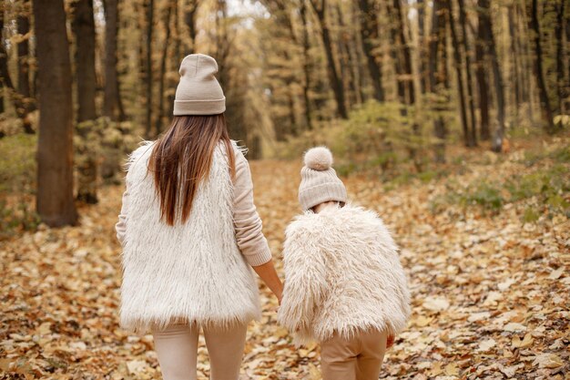 秋の森を歩いている少女と女性の裏側の写真。ブルネットの女性は彼女の娘と遊ぶ。ベージュのセーターを着ている女の子と白い服を着ている母親。