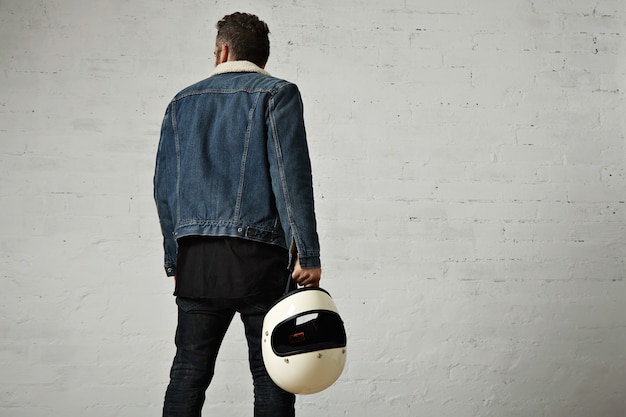 Вид сзади молодой мотоциклист в короткой джинсовой куртке и черной пустой рубашке на хенли, уходит и держит старинный бежевый мотоциклетный шлем, изолированный в центре белой кирпичной стены
