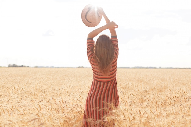 Backview of slender well shaped woman holding her arms up, having straw hat in one hand, standing in front of Sun with pleasure in middle of wheat field, enjoying summer holidays at rural area.