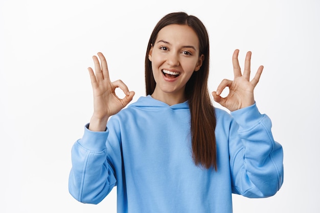 Not bad, congrats. Smiling brunette 20s woman, showing okay OK signs and looking satisfied, praise great choice, good work, give approval, complimenting, standing over white background.