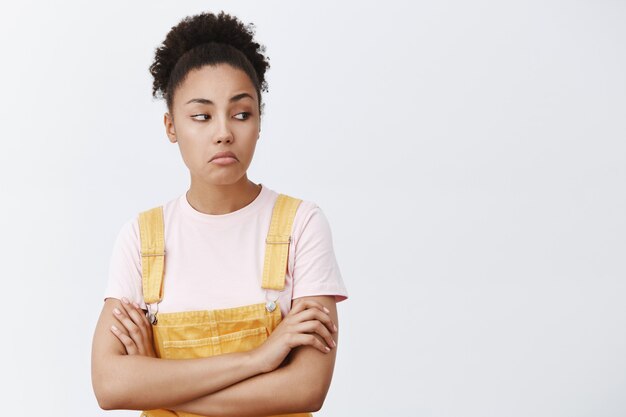 Not bad, nice shoes, want them too. Confident cool African American woman in yellow overalls, pursing lips and gazing right, holding hands crossed on chest, liking something, checking it out