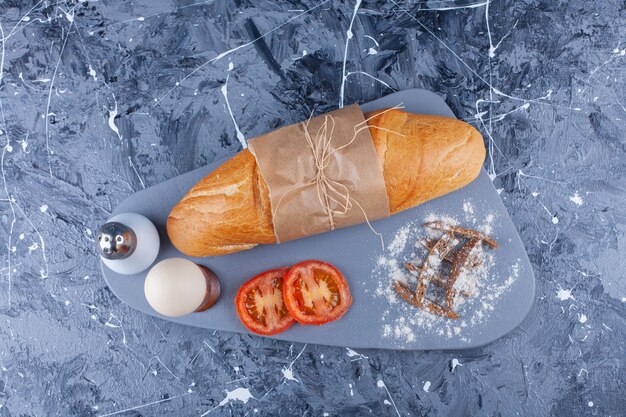 Baguette bread, sliced vegetables, boiled egg on board on blue.