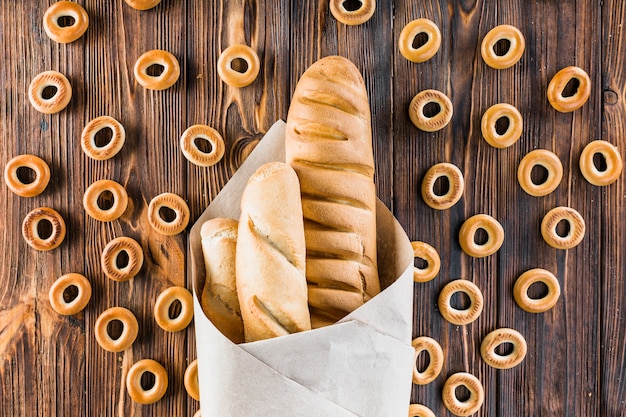 Baguettes wrapped in paper surrounded with bagels on wooden background